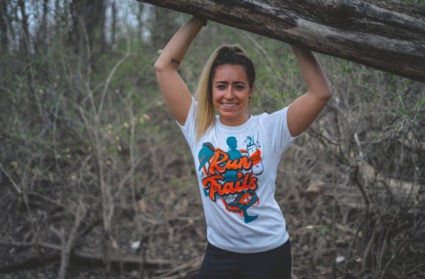 woman with arms above head on a tree limb wearing run trails tee